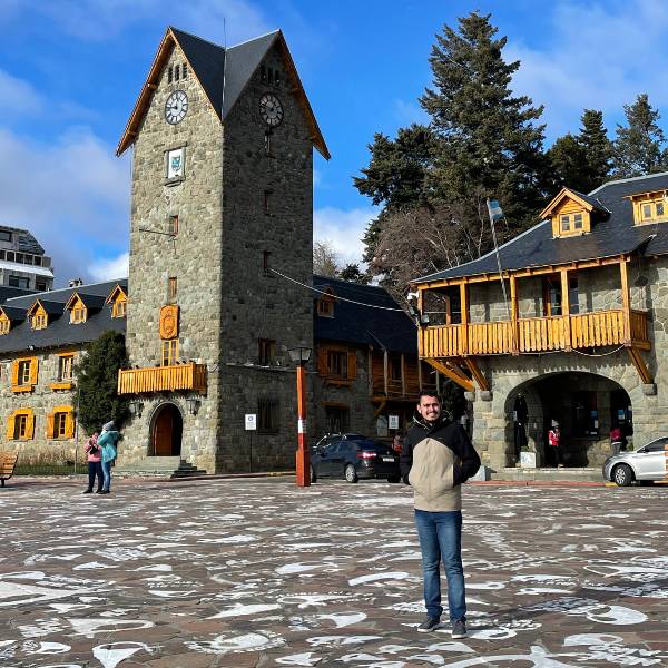 Centro Cívico de Bariloche em dia ensolarado de inverno, sem neve, com pessoas agasalhadas passeando pela praça histórica