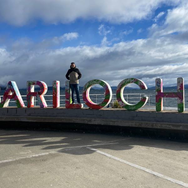 Centro Cívico de Bariloche em dia ensolarado de inverno, sem neve, com pessoas agasalhadas passeando pela praça histórica
