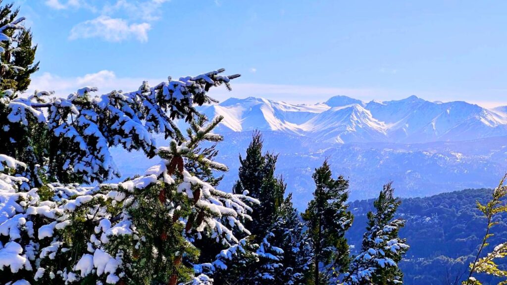 Paisagem de Bariloche coberta de neve, com montanhas ao fundo e um lago cristalino refletindo o céu azul em um dia ensolarado de inverno